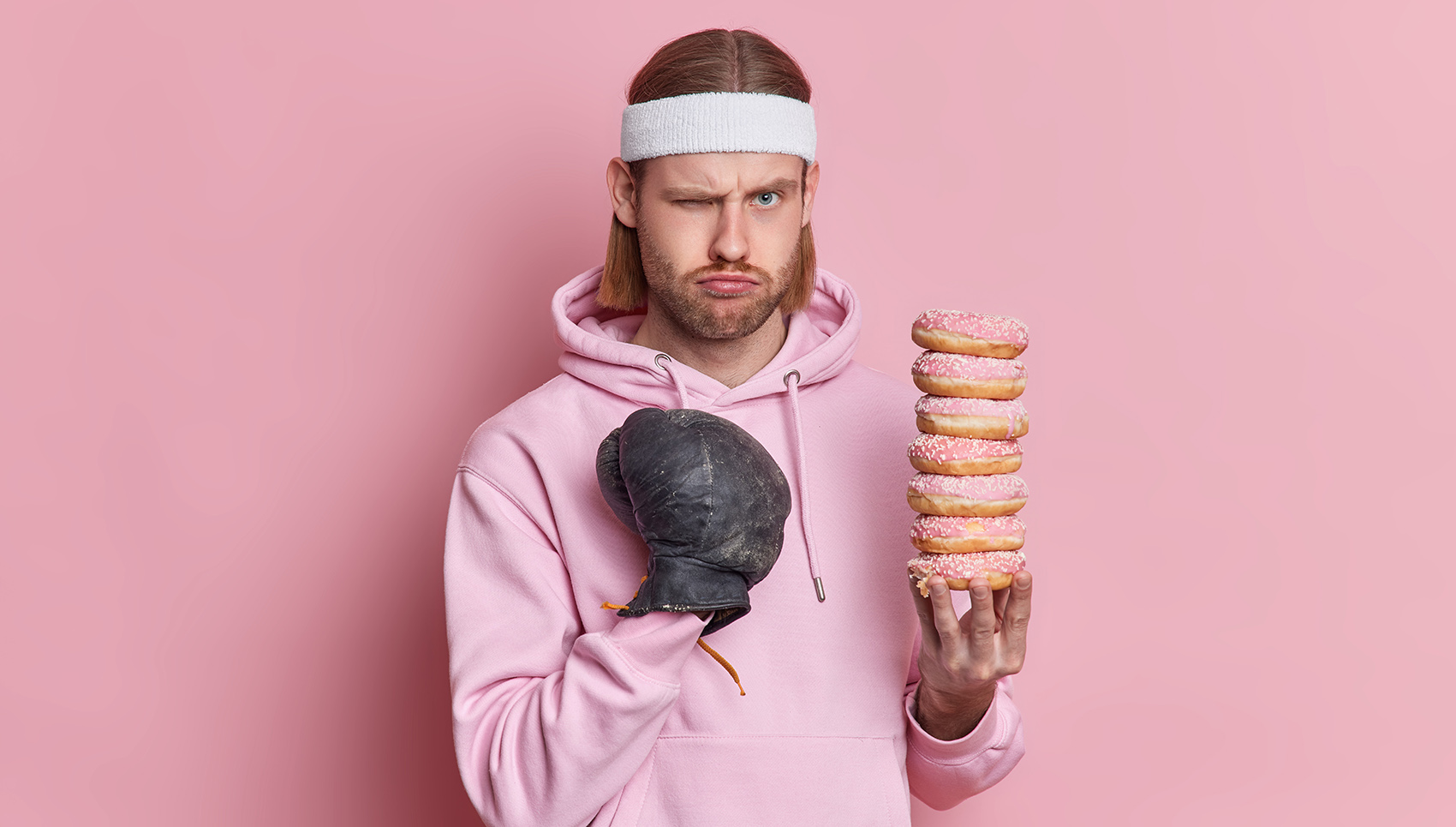 Serious displeased European male boxer makes fist punch ready for fight wears sportclothes holds doughnuts dressed in sweatshirt isolated over pink background. Be strong and healthy concept.